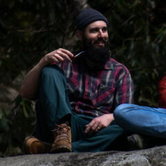 A profile picture of Nick Polk sitting on a rock, wearing a grey beanie, plaid shirt, green pants, and brown boots while holding tobacco pipe